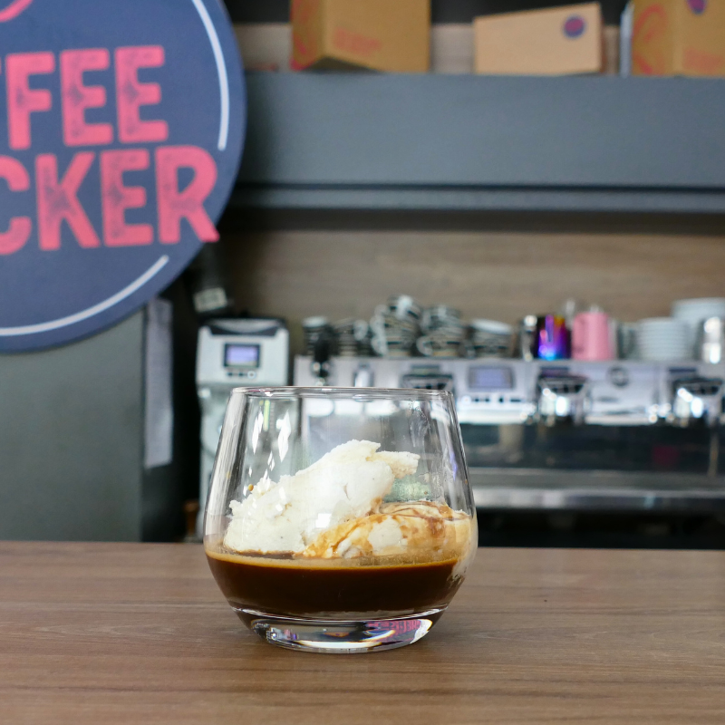Glas mit Affogato al Cafe – Vanilleeis mit Espresso – auf Holztresen vor Siebträgermaschine und Coffeechecker-Logo