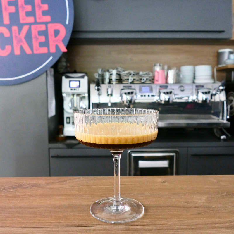 Martini-Cocktailglas mit Cafe Shakerato – geshakter, gekühlter Espresso mit Crema – auf Holztresen vor Siebträgermaschine und Coffeechecker-Logo