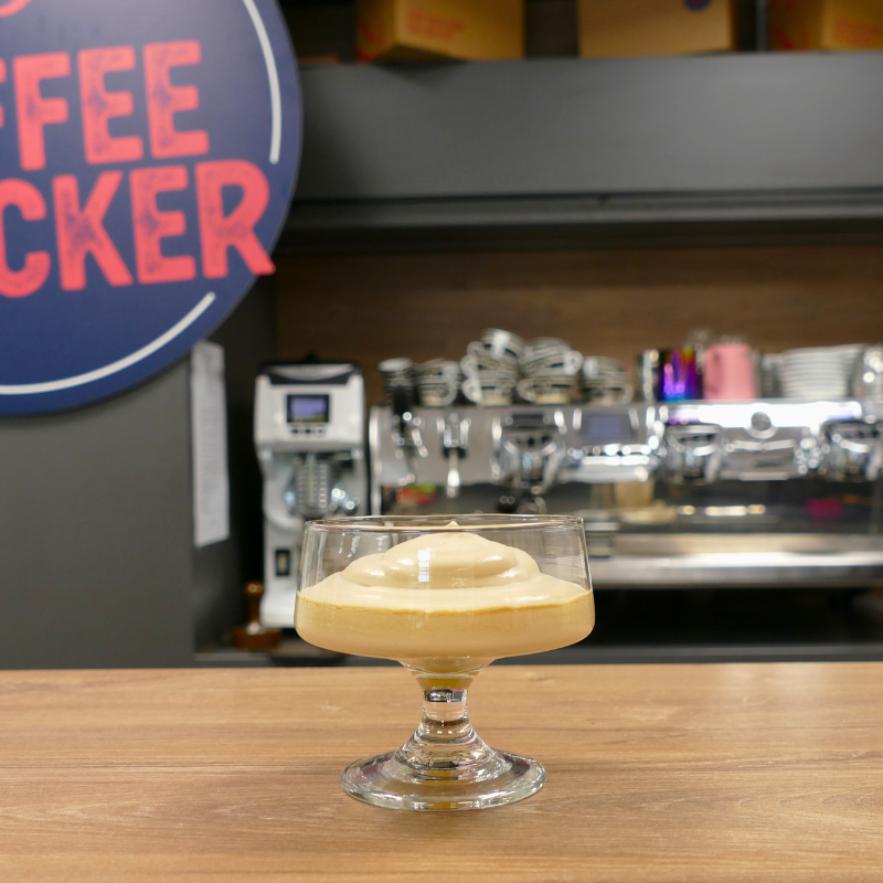 Dessertschale mit Crema di Caffè – aufgeschlagene Kaffeesahne – auf Holztresen vor Siebträgermaschine und Coffeechecker-Logo