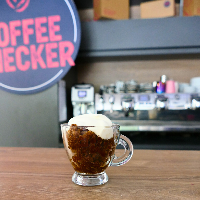 Glastasse mit Granita di Caffè und halbgeschlagener Sahne – auf Holztresen vor Siebträgermaschine und Coffeechecker-Logo
