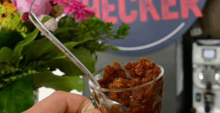 Nahaufnahme einer Hand mit einem Glas voll Granita al Caffè – grobkörniges Kaffeeeis – vor dem Coffeechecker-Logo mit Blumen im Hintergrund