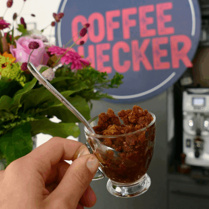Nahaufnahme einer Hand mit einem Glas voll Granita al Caffè – grobkörniges Kaffeeeis – vor dem Coffeechecker-Logo mit Blumen im Hintergrund