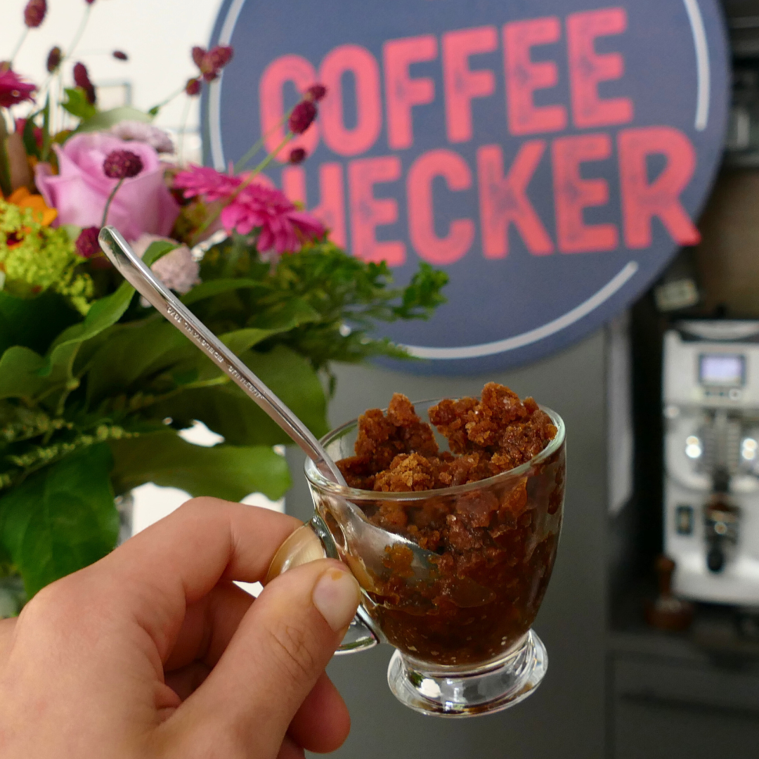 Nahaufnahme einer Hand mit einem Glas voll Granita al Caffè – grobkörniges Kaffeeeis – vor dem Coffeechecker-Logo mit Blumen im Hintergrund